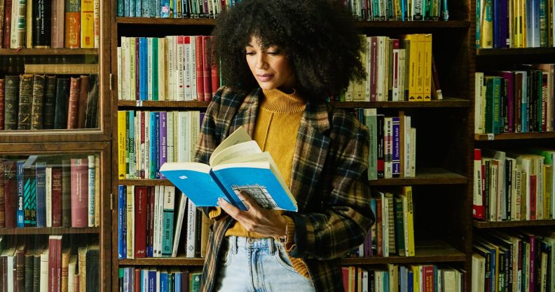 A person in front of an array of books reading from the pages of a paperback.