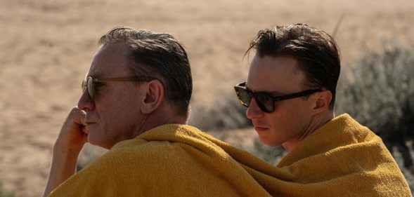 Queer still; Daniel Craig and Drew Starkey sitting together under the same yellow blanket