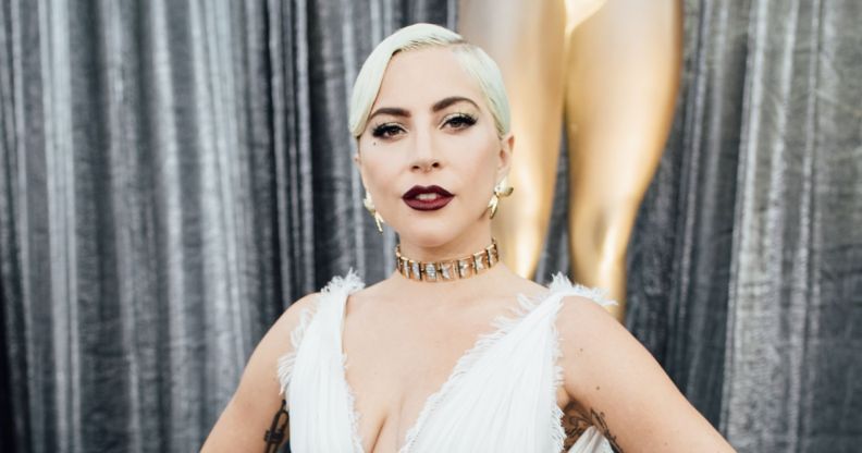 Lady Gaga in a white dress stood against a pair of silver curtains posing.