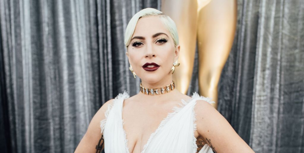 Lady Gaga in a white dress stood against a pair of silver curtains posing.