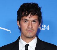 Omar Apollo in a black suit, white shirt and black tie against a blue background at the Queer premiere.