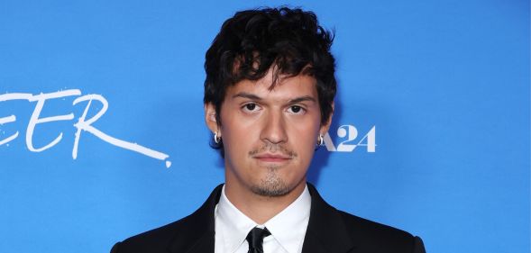 Omar Apollo in a black suit, white shirt and black tie against a blue background at the Queer premiere.