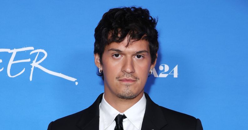 Omar Apollo in a black suit, white shirt and black tie against a blue background at the Queer premiere.