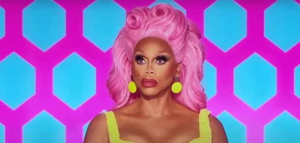 RuPaul in a sleeveless yellow dress and pink wig puckering her lips while sat on the Drag Race All Stars judging panel.