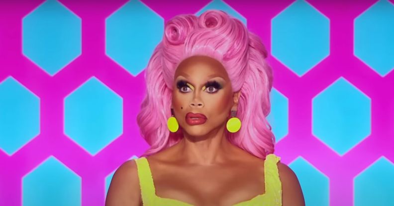 RuPaul in a sleeveless yellow dress and pink wig puckering her lips while sat on the Drag Race All Stars judging panel.