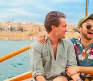 Two men, one in short sleeve colourful t-shirt and the other in a long-sleeve mint shirt, have their arms around each other and are smiling. They are sat on a boat out to sea in Malta.