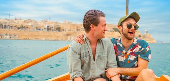 Two men, one in short sleeve colourful t-shirt and the other in a long-sleeve mint shirt, have their arms around each other and are smiling. They are sat on a boat out to sea in Malta.