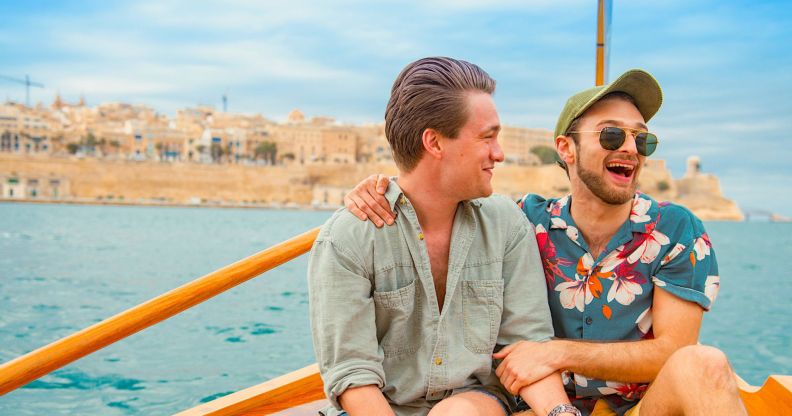 Two men, one in short sleeve colourful t-shirt and the other in a long-sleeve mint shirt, have their arms around each other and are smiling. They are sat on a boat out to sea in Malta.