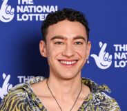 Olly Alexander in a multi-coloured shirt smiling against a blue and white background.