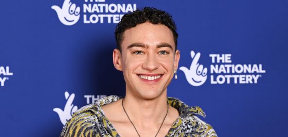 Olly Alexander in a multi-coloured shirt smiling against a blue and white background.