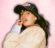 Rebecca Black in a white faux fur jacket and baseball cap, posing with her mouth open and finger and thumb near her face.