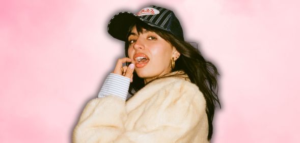 Rebecca Black in a white faux fur jacket and baseball cap, posing with her mouth open and finger and thumb near her face.