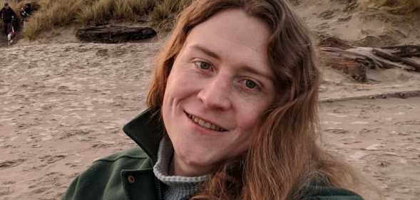 Non-binary American Ryan Castellucci wears a jacket and shirt as they chill on a sandy beach like setting