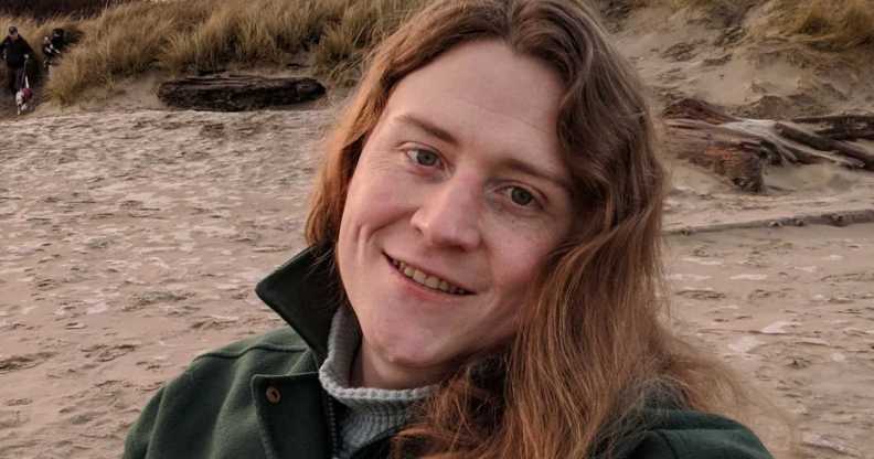 Non-binary American Ryan Castellucci wears a jacket and shirt as they chill on a sandy beach like setting