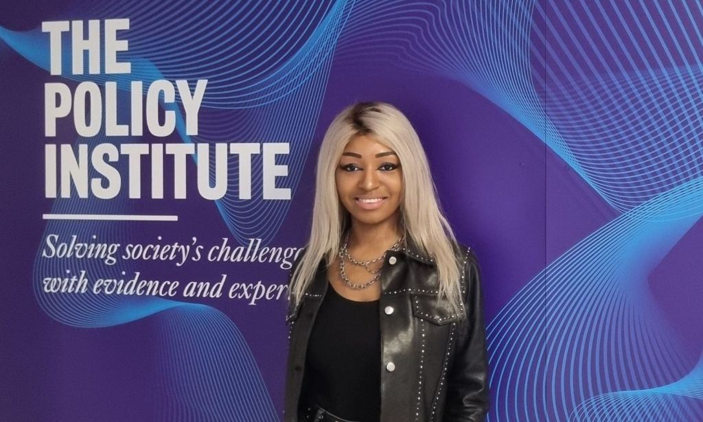 Yasmin Benoit standing next to a wall with text that reads "The Policy Institute: Solving society's challenges with evidence and experts."