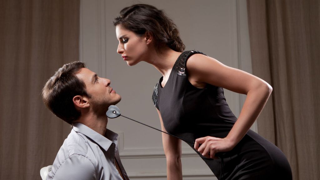 Photo of a man tied to a chair, while a woman with a whip poses above him - they are both fully clothed