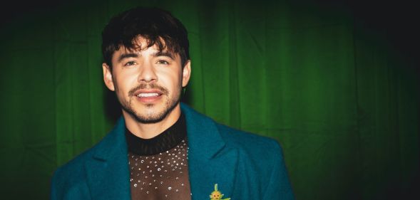 David Archuleta poses for a photoshoot wearing a blue blazer, see through sparkly top and teletubby pin. He is stood against a green curtain