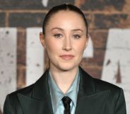 Erin Doherty in a grey shirt, tie and blazer posing against a brick wall