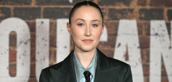 Erin Doherty in a grey shirt, tie and blazer posing against a brick wall