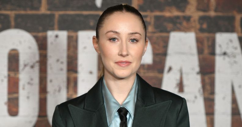 Erin Doherty in a grey shirt, tie and blazer posing against a brick wall