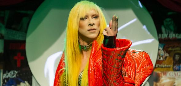 Nick Gaga performing in a blonde wig and red jacket with her fingers raised upwards.