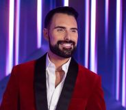 Rylan smiles in a red suit in a promotional image for Doctor Who.