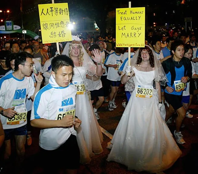 HONG KONG, CHINA: Matt Pearce (L) and Adrian Smith (R) wearing wedding dresses and holding placards run along with thousands of participants in the 10km Men's Open race of Standard Chartered Hong Kong Marathon 2005 in Tsim Tsa Tsui district of Hong Kong, 27 February 2005. Matt and Adrian joined the marathon as a running demonstration calling on the government to allow same-sex-marriage. "We chose the marathon because it is a middle class family event and it's that class of people who are preventing gays the right to marry," said Matt Pearce, protest co-ordinator and spokesman for activist group International Action. Homosexuality was only decriminalised in Hong Kong in 1991 and the age of consent for gays is 21 even though for heterosexuals it is 16. Gay marriage is still banned. Gay and lesbian groups say authorities have hardened their opposition since rule of Hong Kong was transferred in 1997 to China, where homosexuality, though lawful, is stigmatised. AFP PHOTO/TED ALJIBE (Photo credit should read TED ALJIBE/AFP/Getty Images)