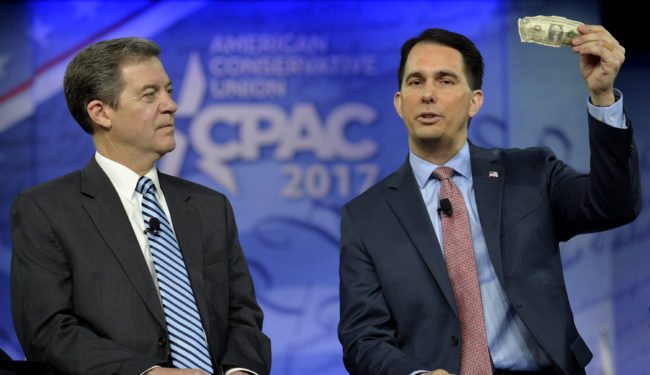 Wisconsin Gov. Scott Walker (R) holds up a US one dollar bill as he makes remarks on taxes as Kansas Gov. Sam Brownback listens during a panel discussion at the Conservative Political Action Conference (CPAC) at National Harbor, Maryland, February 23, 2017.   Politicians, pundits, journalists and celebrities gather for the annual conservative event to hear speakers, network and plan agendas for the new President Trump administration.               / AFP / Mike Theiler        (Photo credit should read MIKE THEILER/AFP/Getty Images)