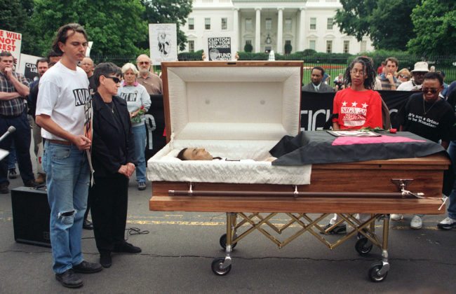 WASHINGTON, UNITED STATES: Aids activists participate in a protest march by opening the casket of AIDS activist Steve Michael as his partner Wayne Turner (L) and his mother Barbara Michael (2nd-L) look on in front of the White House in Washington, DC 04 June 1998. The protesters mourned the death of Michael, the Act Up Washington, DC founder, who died as a result from AIDS 25 May. The group denounced US President Bill Clinton's campaign promises to find a cure for the disease. AFP PHOTO/Jamal A. WILSON (Photo credit should read JAMAL A. WILSON/AFP/Getty Images)
