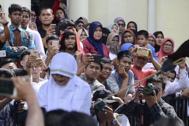 TOPSHOT - EDITORS NOTE: Graphic content / Onlookers watch as a member of Indonesia's Sharia police (at R-outside frame) whips a non-Muslim woman (bottom L) for trading alcohol during a public caning ceremony outside a mosque in Banda Aceh, the capital of Aceh province on July 13, 2018. - A gay couple was also publicly whipped in Indonesia's conservative Aceh province on July 13, despite an earlier pledge by officials to stop the punishment after it drew international criticism. (Photo by CHAIDEER MAHYUDDIN / AFP) (Photo credit should read CHAIDEER MAHYUDDIN/AFP/Getty Images)