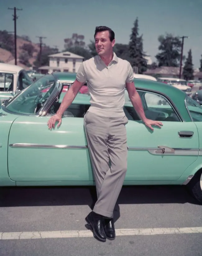 American actor Rock Hudon (1925 - 1985) leans against a sea foam green car in a parking lot, 1950s